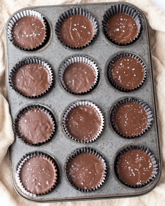 Keto Peanut Butter Chocolate Bites - Overhead view of a muffin tin with 12 shiny dark chocolate cups in brown liners, sprinkled with sea salt on beige cloth
