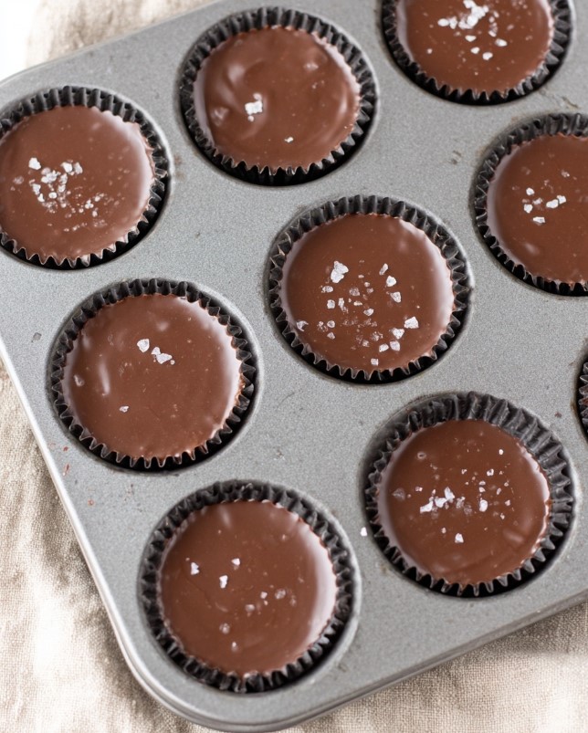 Low Carb Peanut Butter Chocolate Bites - Angled view of muffin tin with 12 chocolate cups showing glossy finish and salt topping, warm lighting