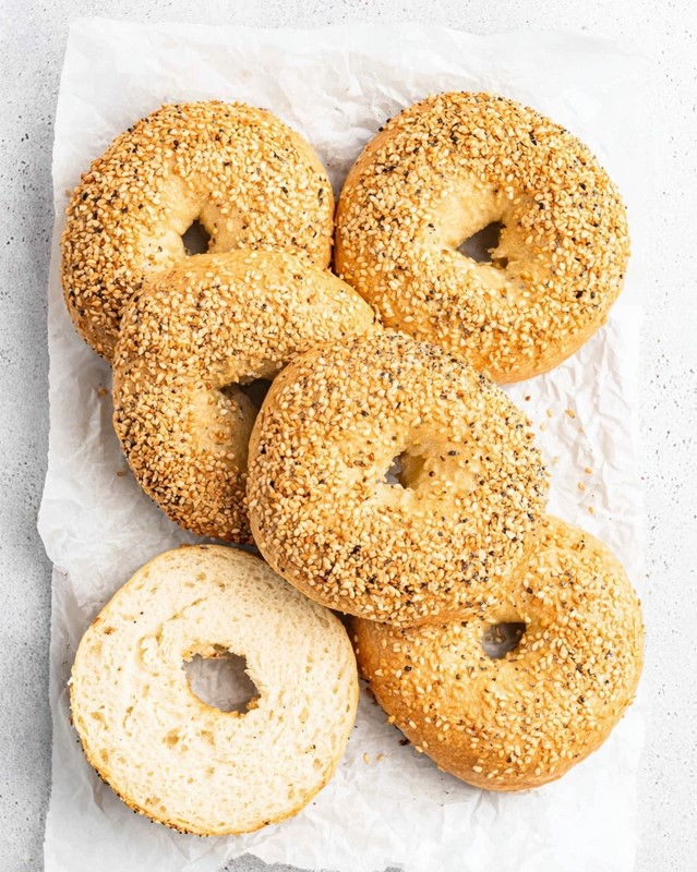 A collection of baked and toasted bagels arranged on a surface, with one cut in half to show the perfectly developed crumb inside, ready for serving.