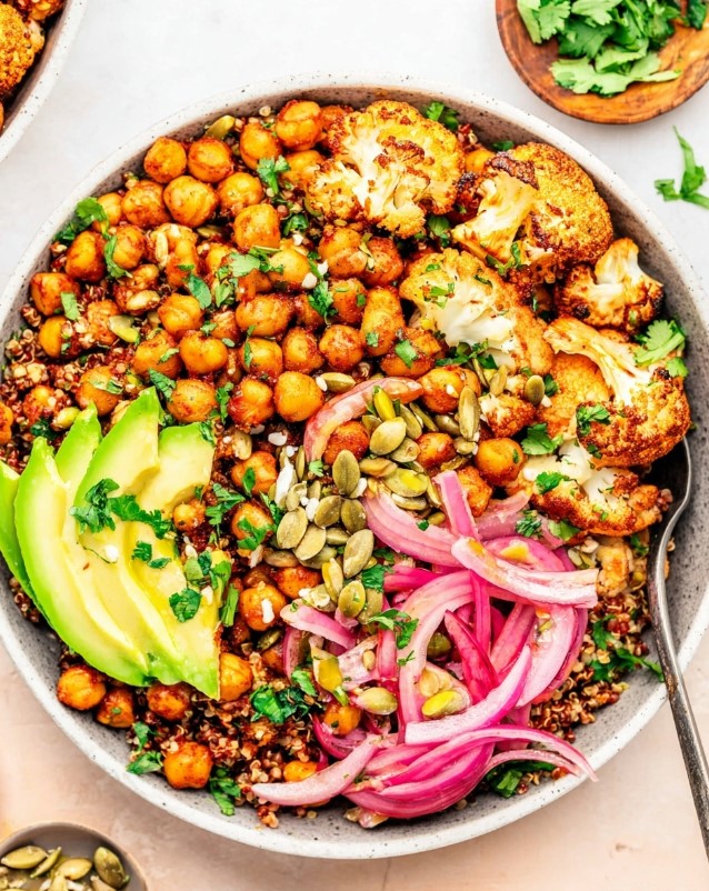 A colorful bowl filled with fluffy quinoa, golden roasted cauliflower, crispy chickpeas, sliced avocado, and pickled red onions, garnished with fresh cilantro and seeds.