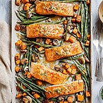 A complete sheet pan dinner featuring roasted salmon fillets glazed in tahini, surrounded by golden sweet potato cubes, roasted mushrooms, and vibrant green beans, ready to serve.