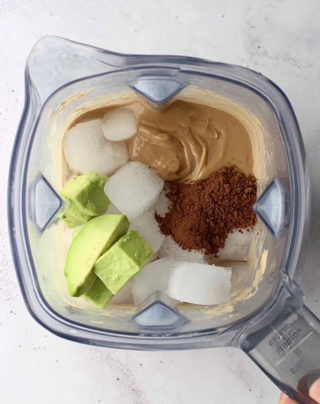 Ingredients like fresh avocado, cocoa powder, and ice layered in a blender, ready to mix.