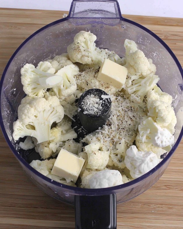 Raw cauliflower florets placed in a food processor with cubes of butter, cream cheese, and seasonings before blending.
