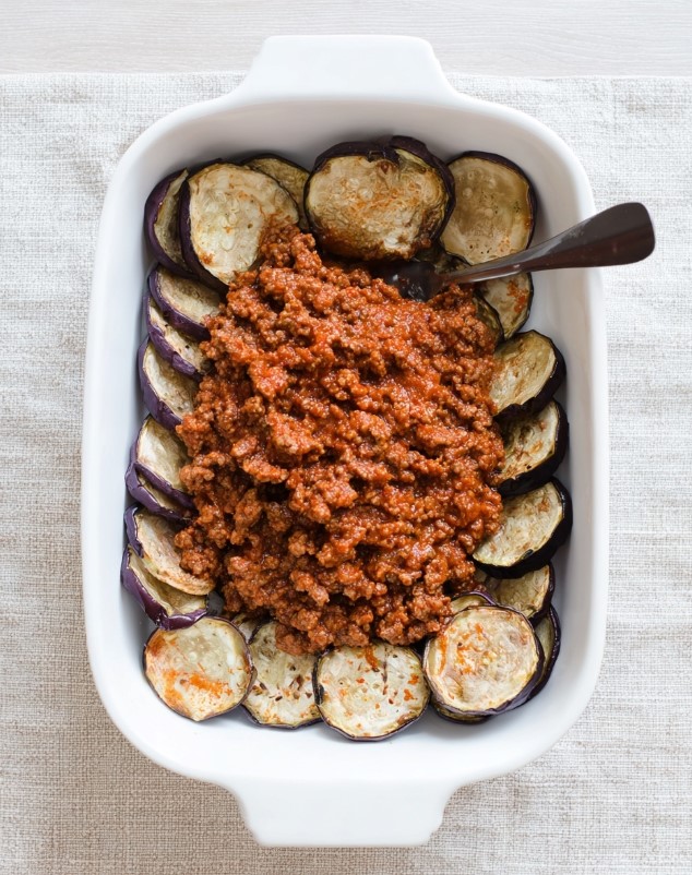 The casserole dish during assembly, showing a layer of roasted eggplant slices being topped with the thick meat sauce.