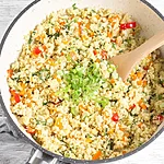 Better-Than-Takeout Keto Cauliflower Fried Rice for Busy Weeknights A close-up, top-down view of a plate of golden fried cauliflower rice mixed with bright red peppers and yellow egg bits, freshly garnished with chopped green scallions.