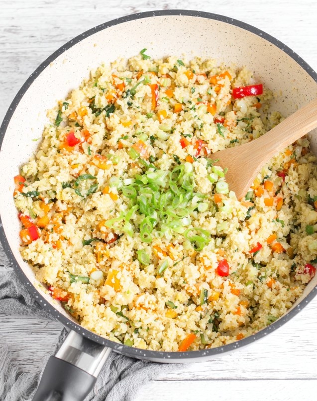 A close-up, top-down view of a plate of golden fried cauliflower rice mixed with bright red peppers and yellow egg bits, freshly garnished with chopped green scallions.
