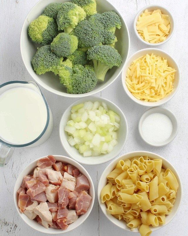 Cheesy Chicken and Bacon Pasta Bake Ingredients - raw broccoli florets, dry pasta, shredded cheese, milk, chopped onion, and diced chicken and bacon arranged on a table