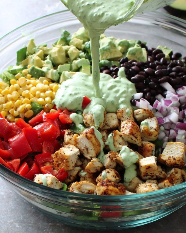 A bowl containing separate piles of diced chicken, avocado, black beans, corn, red pepper, and onion, with a bright green sauce being poured over the top