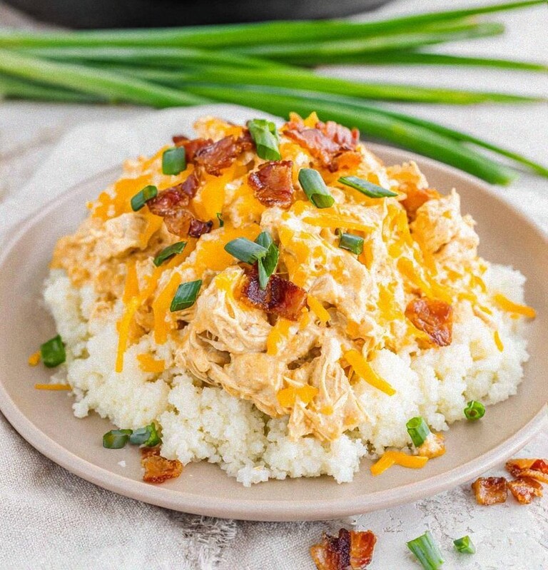 Plated slow cooker keto crack chicken served over a bed of cauliflower rice and topped with melted cheddar cheese, chopped bacon, and green onions