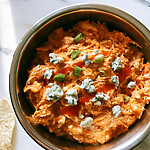 Creamy orange Instant Pot buffalo chicken dip in a metal bowl topped with blue cheese, scallions, and extra hot sauce