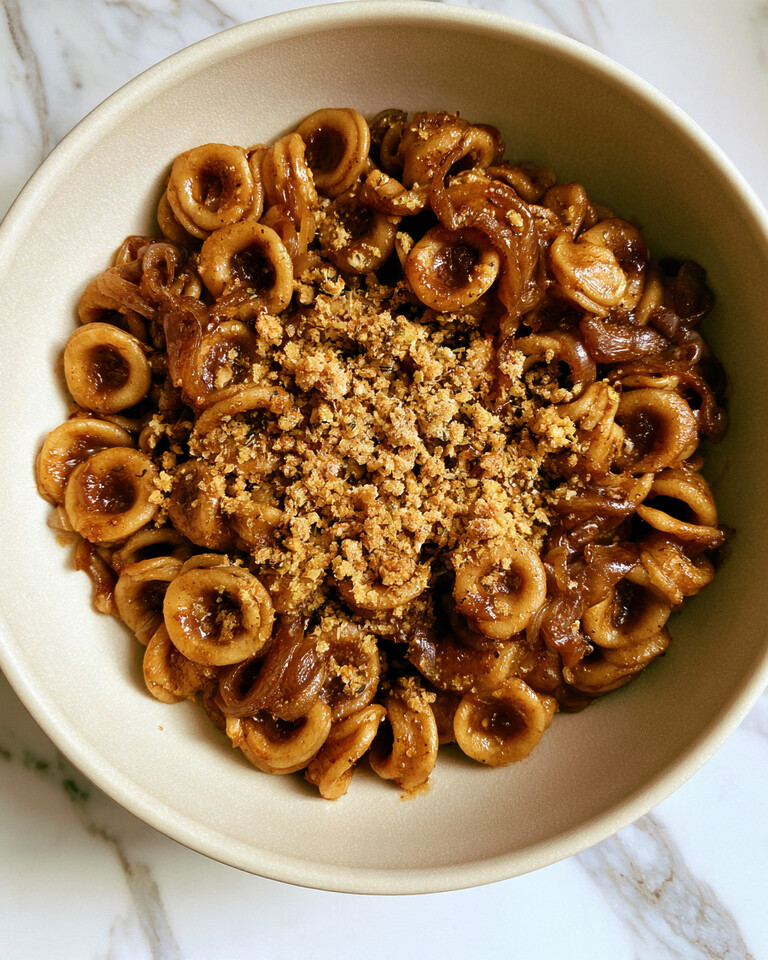Light ceramic bowl of French onion pasta with glossy orecchiette shells, caramelized onions, and toasted garlic breadcrumbs in the center