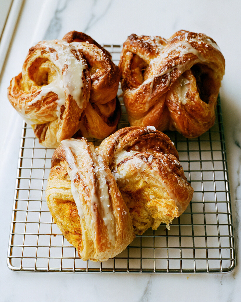 Freshly baked golden pumpkin cruffins on a metal cooling rack with white maple cream cheese glaze flowing over the layers