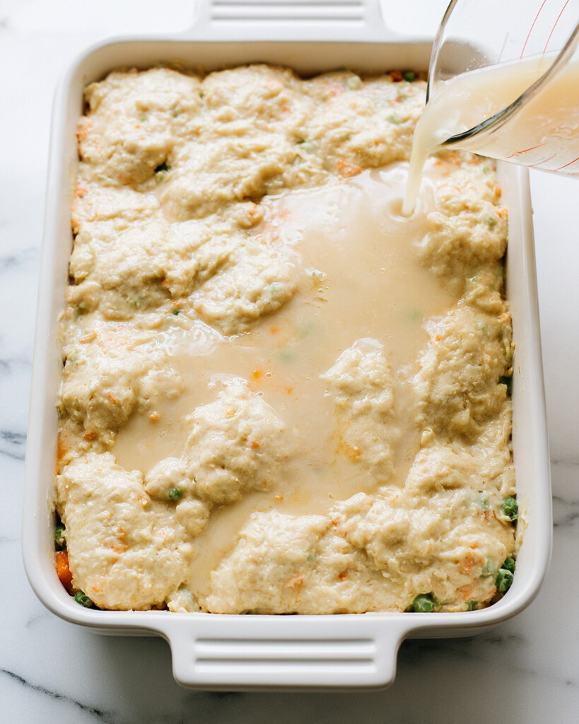 Light chicken broth and soup mixture being poured over thick cheddar biscuit batter in a baking dish