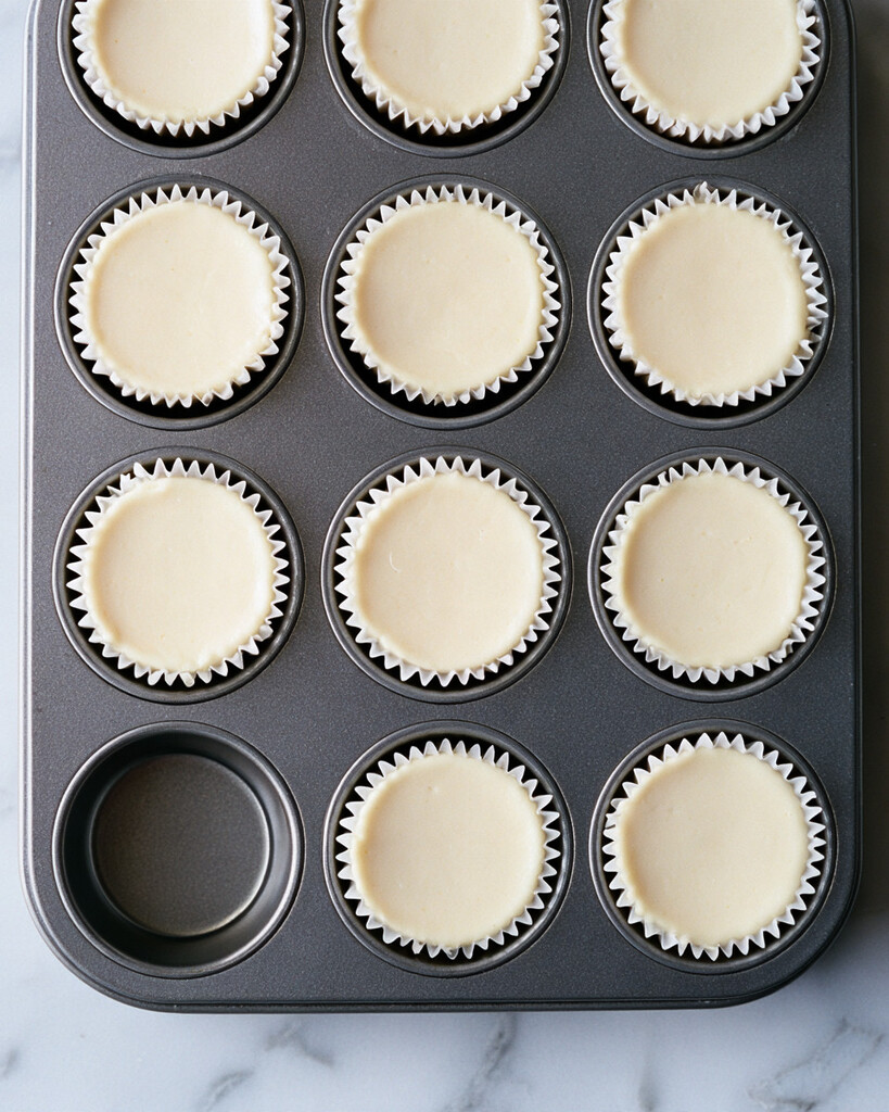 Smooth cheesecake batter filling paper cups over baked cookie crusts in a muffin pan